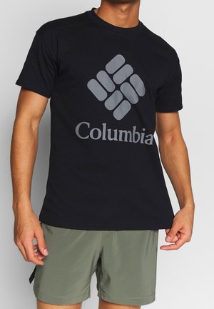 Mand iført en sort Columbia T-shirt med stort logo og olivengrønne shorts, stående mod en ensfarvet lys baggrund.