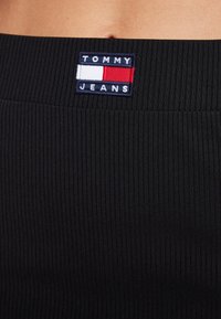 Černý žebrovaný pas s vyšívaným logem "TOMMY JEANS" v modré, bílé a červené barvě. Materiál vykazuje viditelnou texturu a strukturovaný design.