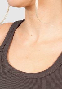 Brun rippad linne med rundad halsringning och synliga sömdetaljer, bärs mot en neutral hudton i en enkel, ljus bakgrund.