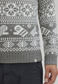 Grå strikket sweater med hvide snefnug og geometriske mønstre. Ribbede kanter og ærmer, med en lille mærketag nederst til venstre.