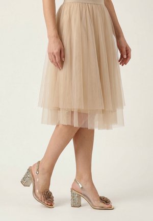 Kobieta ubrana w beżową, warstwową tiulową spódnicę oraz przezroczyste sandały na obcasie ozdobione materiałowymi różami i koralikami.