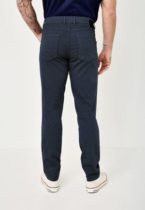 Mann, der dunkelblaue Hose mit geradem Bein, weiße Turnschuhe und ein marineblaues Hemd trägt, von hinten gezeigt mit sichtbaren tätowierten Armen.