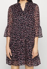 Svart blommig klänning med V-ringning, genomskinliga trekvartsärmar och trumpetärmar. Har små rosa och röda blommor samt en flerradig kjol.
