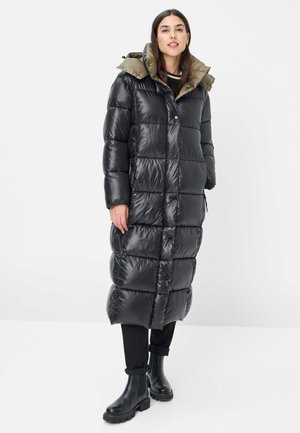 Frau trägt einen langen schwarzen Steppmantel mit grünem Kapuzenfutter, schwarze Hose und schwarze Stiefeletten, steht vor einem schlichten weißen Hintergrund.
