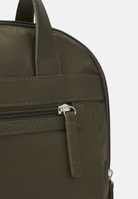 Primer plano de la esquina de una mochila verde oliva, mostrando la tela cosida, el zipper con una lengüeta de cuero y un pequeño asa en un fondo blanco.
