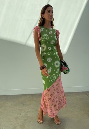 Kvinne iført en grønn og rosa mønstret maxikjole med volangerm, holder en flerfarget clutch og står i et minimalistisk rom.