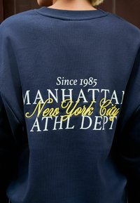 Sweat-shirt navy avec des lettres blanches et jaunes : "Depuis 1985 Manhattan New York City ATHL DEPT" sur le dos d'une personne.