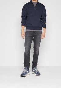 Marinblå zip-up huvtröja med framficka, kombinerad med mörkgrå jeans och marinblå sneakers med vita detaljer och en logotyp.