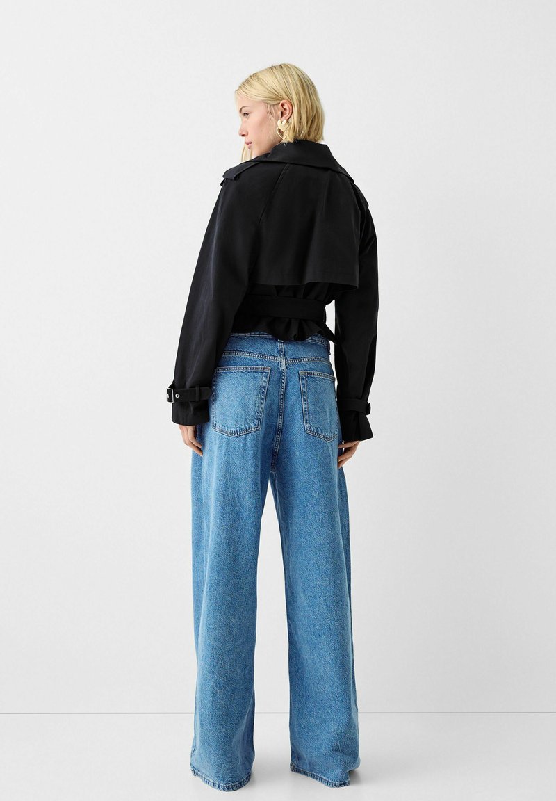 Donna che indossa una giacca nera cropped con dettagli a fibbia e jeans a gamba larga blu, in piedi contro uno sfondo bianco semplice, rivolta di spalle.