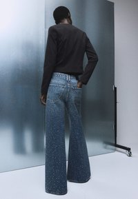 Jeans in denim a gamba larga con una vestibilità blu chiaro, caratterizzati da un alto punto vita e macchie bianche sparse. Abbinati a un top nero a maniche lunghe corto.