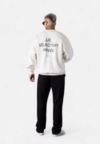White sweatshirt with "LA SÉLECTION PRIVÉE" printed on the back, paired with black pants. Model is wearing sunglasses.
