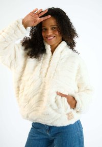 Giacca a pullover fuzzy di colore crema realizzata in materiale peloso, con colletto alto e tasche laterali. Presenta motivi testurizzati e un'etichetta con il logo.