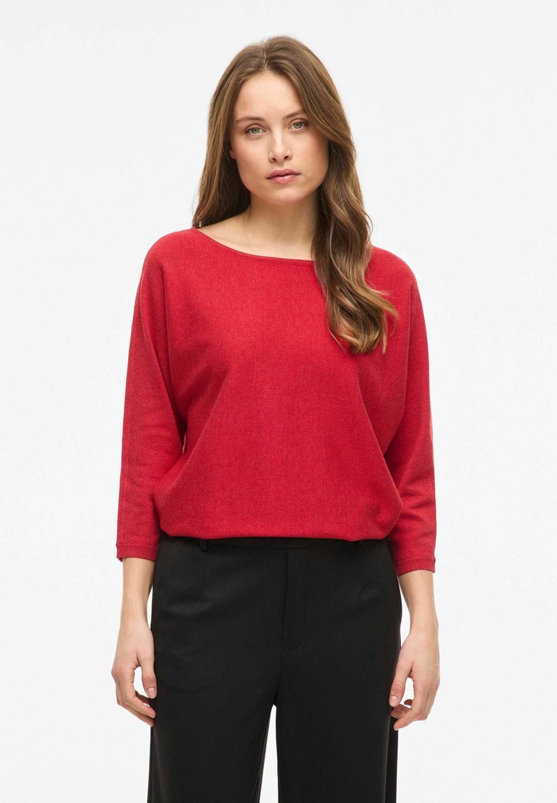 Femme aux longs cheveux bruns ondulés portant un pull rouge à encolure bateau et un pantalon noir, debout devant un fond blanc.