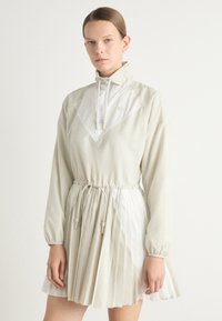 Lätt, krämfärgad jackklänning med hög krage, dragsko i midjan och plisserad kjol. Har kontrasterande paneler och mjuk textur.