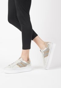 Sneaker grigio chiaro e crema con dettagli metallici, pannelli in tessuto testurizzato, suola bianca spessa e occhielli decorativi.