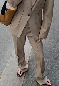 Beżowy garnitur dopasowany z pionowymi przeszyciami, w połączeniu z czarnymi sandałami. Brązowa torebka z zamszu ma zaokrąglony kształt i pasek na ramię.