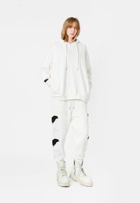 Sudadera blanca de gran tamaño con bolsillo frontal, combinada con pantalones blancos ajustados que presentan bordado de alas negras. Llevada con botas blancas robustas.
