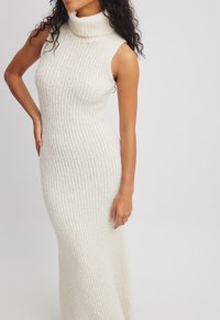 Rebrast obleka iz bež pletenine z visokim turtleneck-om in brezrokavnim dizajnom, ki ima oprijeto silhueto in mehak teksturo.