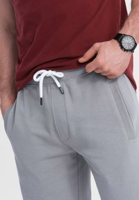 Pantalones de chándal grises con un cordón blanco, con bolsillos laterales y una textura suave, combinados con una camiseta color burdeos y un reloj negro.