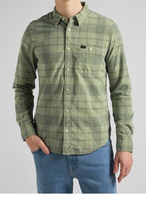 Mann trägt grünes kariertes Hemd mit Brusttasche, auf der "Lee" steht, und blaue Jeans, steht mit einer Hand in der Tasche.