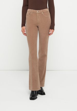 Femeie purtând pantaloni evazați bej din catifea de tip cord, bluză maro cu mâneci lungi cu dungi și ghete negre până la gleznă pe fundal alb.