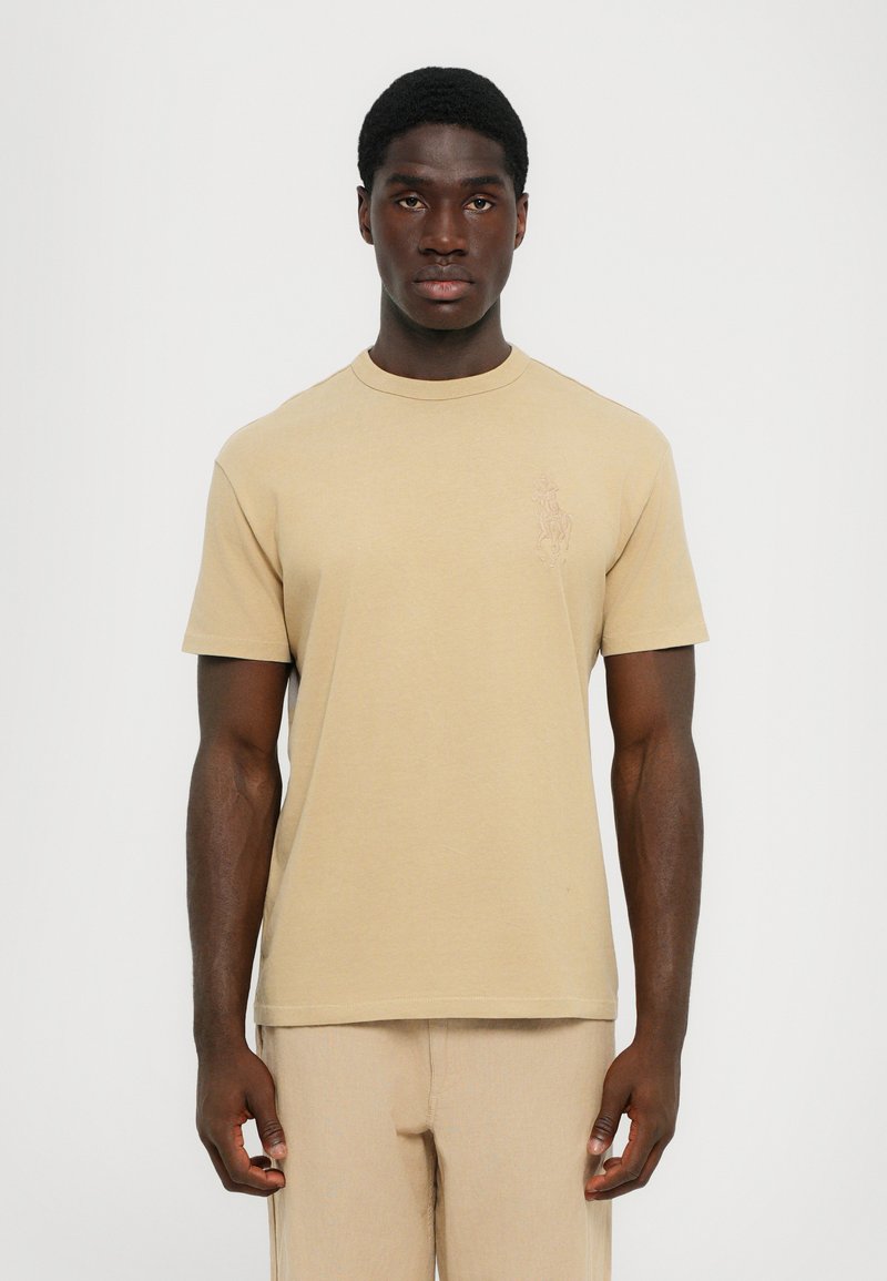 Beige kortärmad t-shirt med rund halsringning och diskret broderad design på vänster bröst. Tillverkad av mjuk bomullstyg.