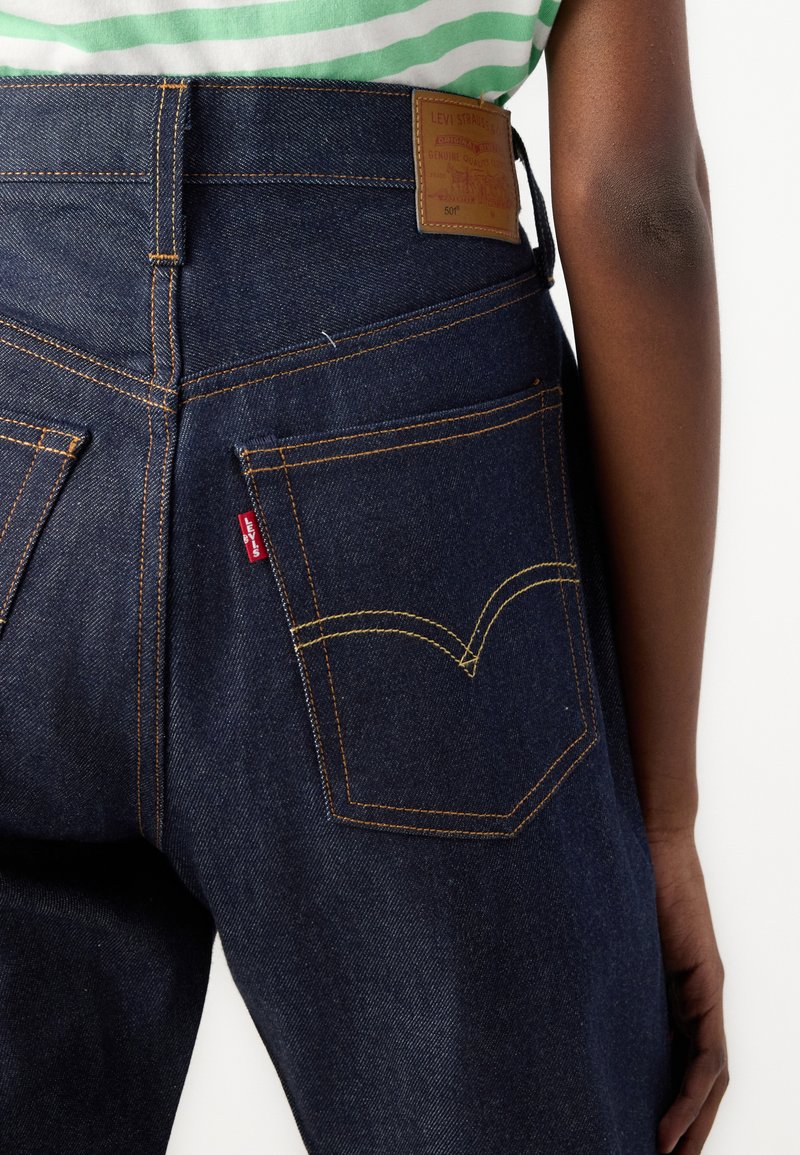 Jeans de mezclilla azul oscuro con una cintura alta, costuras amarillas, dos bolsillos traseros con un diseño de costura curva característico y una etiqueta de cuero.
