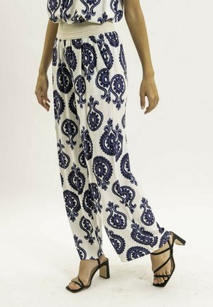 Femme portant un pantalon blanc à jambes larges avec des motifs paisley bleus et des sandales à talons hauts noires, debout devant un fond uni.