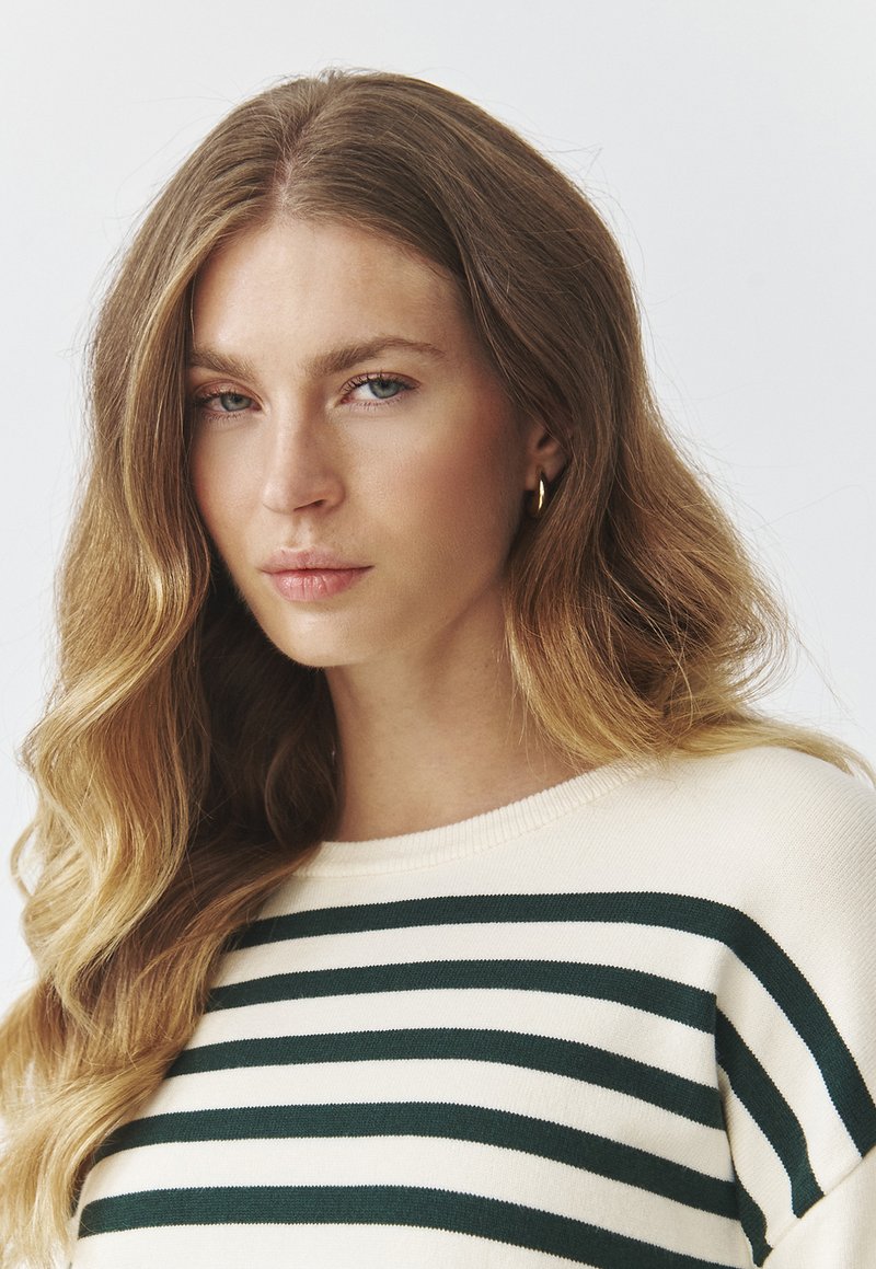 Kremowy sweter z ciemnozielonymi poziomymi paskami, wykonany z miękkiego materiału. Dekolt jest okrągły, a struktura wydaje się gładka i przytulna.