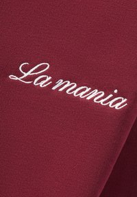 Bordo audinys su tekstūru, kuriame yra baltais kursyviniais rašmenimis išsiuvinėtas tekstas "La mania". Detalus artimas vaizdas