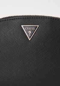 Pochette in tessuto nero testurizzato con logo triangolare in metallo con la scritta "GUESS" e un punto interrogativo. Accenti metallici dorati lungo la cerniera.