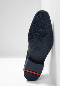 Semelle de chaussure noire avec texture en nid d'abeille, présentant un accent rouge au talon. Bord supérieur lisse avec une légère courbure.