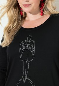 Blondýna s červenými náušnicemi a černým topem s obrysem ženy v blejzrových šatech a podpatcích zdobeném kamínky.