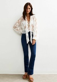 Doorzichtig wit bloesje met bloemenborduursel, gestrikt bij de hals, gecombineerd met donkerblauwe flared jeans en bruine platform sandalen.