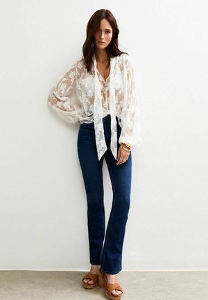 Blusa bianca trasparente con ricami floreali, legata al collo, abbinata a jeans flared blu scuro e sandali platform marroni.
