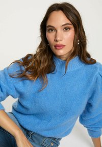 Pullover blu a maglia con collo alto, maniche a palloncino e una trama morbida. Abbinato a jeans, il colore è vibrante e ricco.