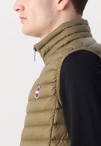 Colete acolchoado verde-oliva com colarinho alto, costura vertical e um emblema redondo. Usado sobre um suéter preto. Fecho com zipper.