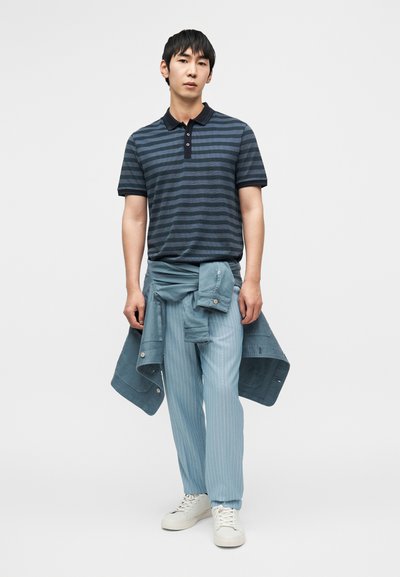 Ριγέ μπλε ναυτικό και μπλε πόλο μπλουζάκι, συνδυασμένο με ανοιχτό μπλε παντελόνι με λεπτές ρίγες· casual μπουφάν δεμένο γύρω από τη μέση, λευκά αθλητικά παπούτσια.