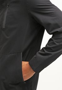 Osoba ubrana w czarną kurtkę z zamykaną kieszenią na piersi, z ręką włożoną do bocznej kieszeni, widziana od połowy tułowia do ramienia.