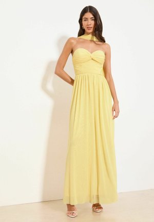 Vrouw draagt een strapless gele maxi-jurk met een geplooide lijfje en een doorschijnende sjaaldetail, staande tegen een effen witte achtergrond.