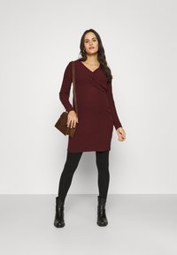 Bordeaux ribbel jurk met lange mouwen, gedraaide voorkant, gecombineerd met zwarte leggings en enkellaarsjes, met een bruine tas.