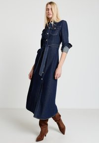 Jeanshemdkleid in Dunkelblau mit tailliertem Gürtel, aufgesetzten Taschen mit Knöpfen und hochgekrempelten Ärmeln. Kombiniert mit braunen Wildleder-Stiefeln mit Absatz.