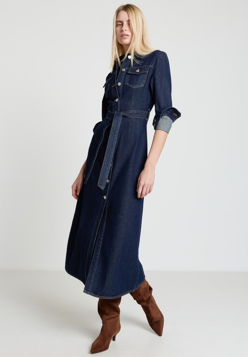 Jeanshemdkleid in Dunkelblau mit tailliertem Gürtel, aufgesetzten Taschen mit Knöpfen und hochgekrempelten Ärmeln. Kombiniert mit braunen Wildleder-Stiefeln mit Absatz.