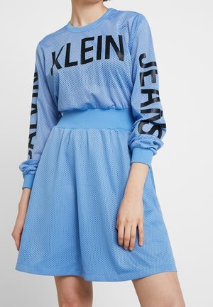 Jasnoniebieska sukienka z siateczki z długimi rękawami, z czarnym napisem "KLEIN" na piersi i "JEANS" na rękawie, z elastycznym paskiem w talii, noszona przez stojącą osobę.