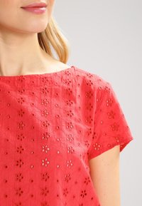Červený vyšívaný top s květinovými výřezy, krátkými rukávy a kulatým výstřihem, vyrobený z lehkého materiálu.
