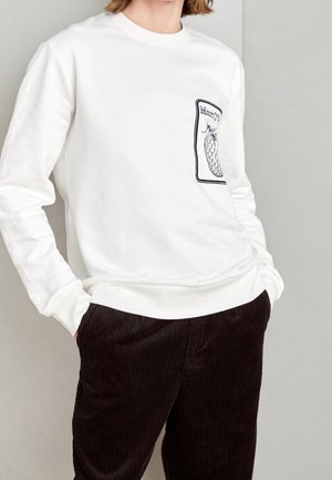 Vit sweatshirt med långa ärmar, med en texturerad ananasdesign i svart på vänster bröstficka. Mörkbruna corduroybyxor.