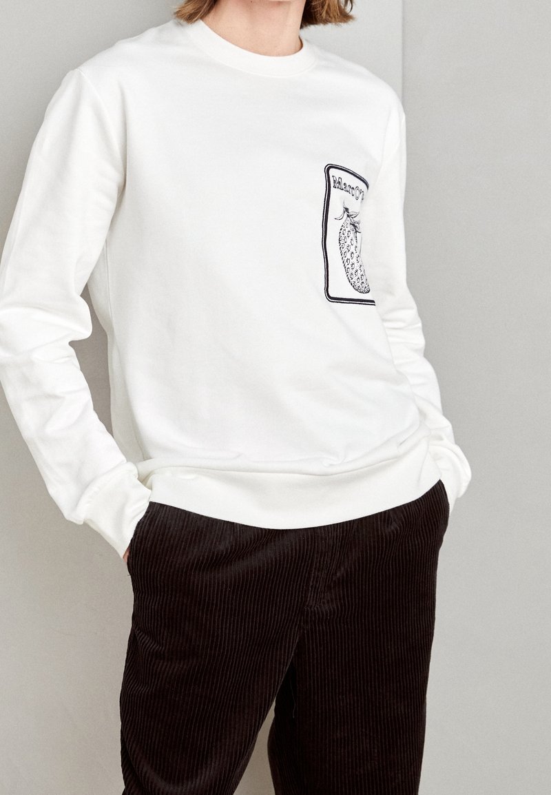 Vit sweatshirt med långa ärmar, med en texturerad ananasdesign i svart på vänster bröstficka. Mörkbruna corduroybyxor.