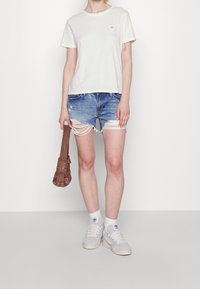 Nainen, joka pitää yllään valkoista T-paitaa, revittyjä farkkushortseja, valkoisia Adidas-tennareita ja pitelee pientä ruskeaa käsilaukkua tasaisella taustalla.