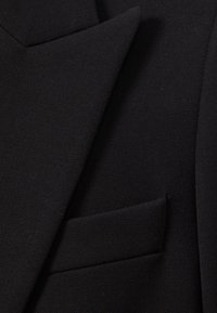 Primo piano di una giacca da completo nera strutturata che mostra il rever e una tasca a filetto sul davanti.