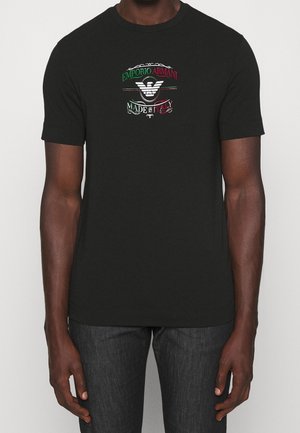 Mand iført en sort T-shirt med korte ærmer med et centralt Emporio Armani-logo og teksten "Made in Italy" i rødt, hvidt og grønt.